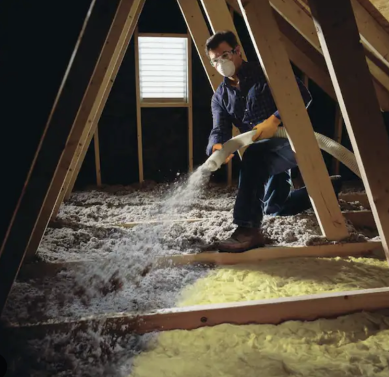 man with a mask and protective eye gear kneeling down and blowing cellulose insulation through a hose in an attic