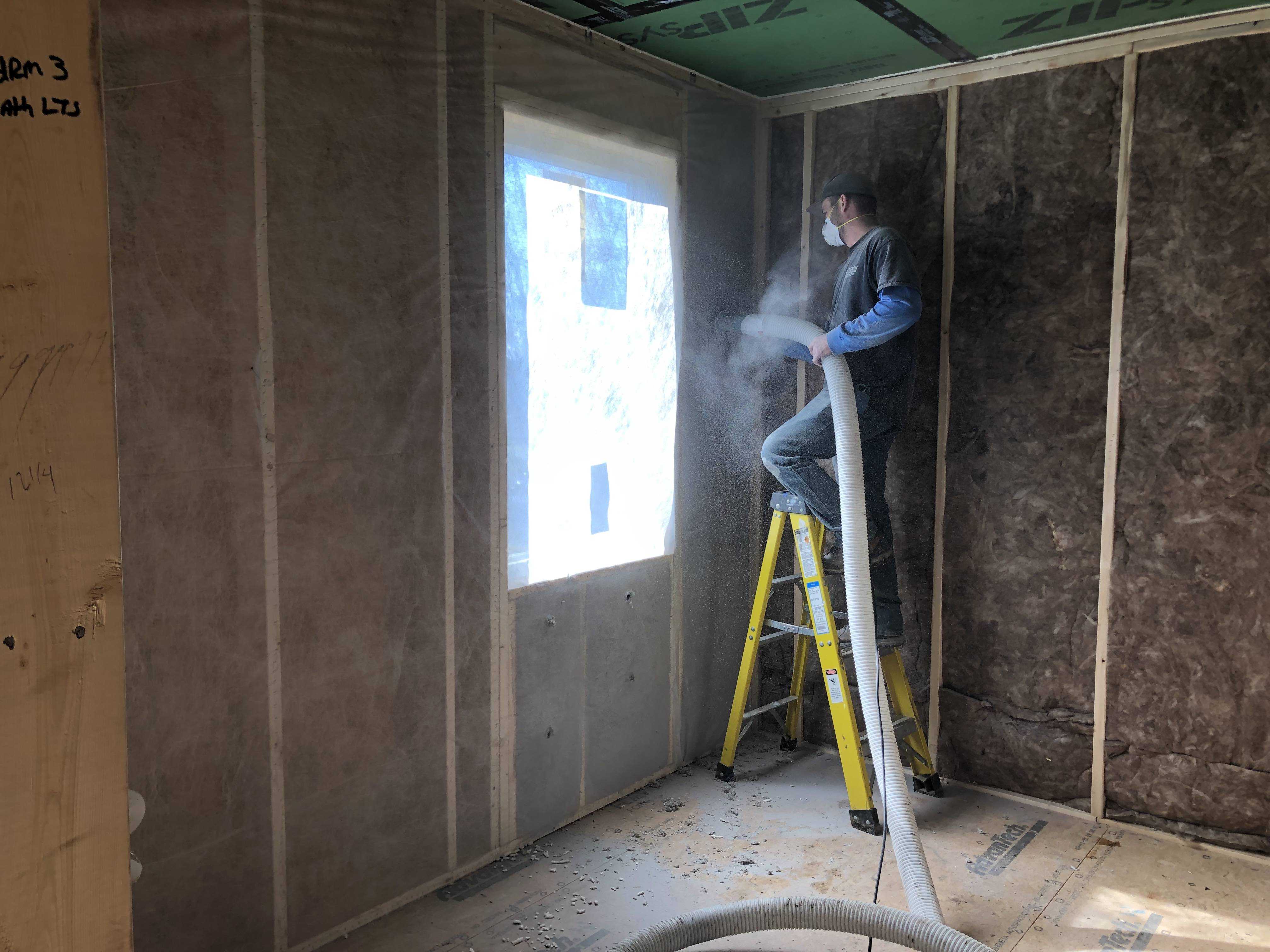 man on a ladder inside an unfinished room blowing insulation in the walls